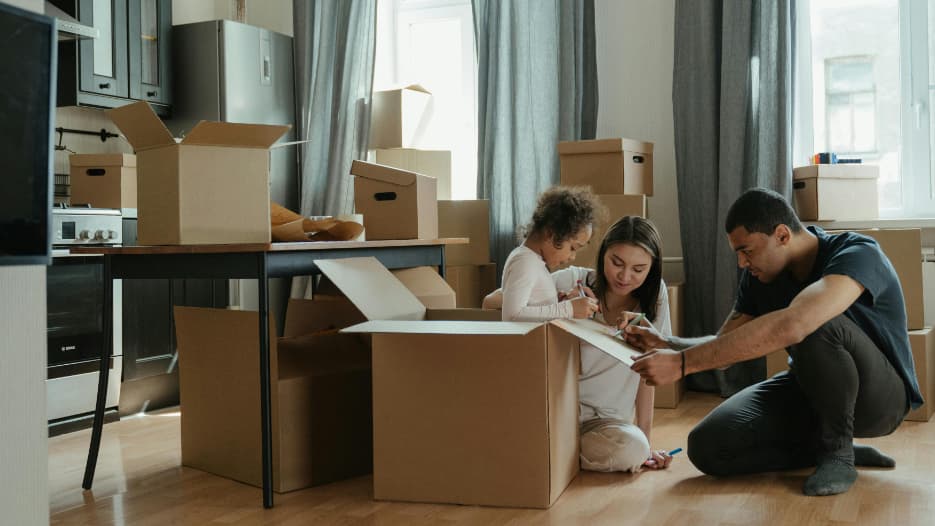 Junge Familie mit Kind sitzt zwischen Umzugskartons auf dem Boden und beschriftet gemeinsam einen Karton.